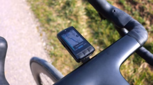 Wahoo ELEMNT BOLT Fahrradcomputer am Cockpit eines Rennrads, aufgenommen während einer Fahrt auf einem Weg neben einer Wiese.