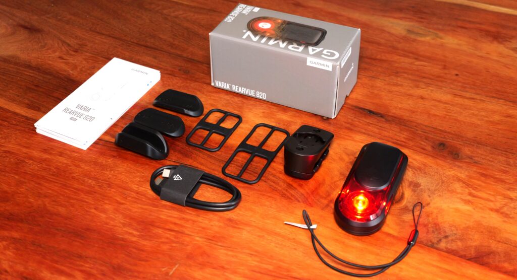 Lieferumfang des Garmin RearVue 820 auf einem Holztisch: Rücklicht, Halterung, Gummibänder, Abstandshalter, USB-Kabel, Sicherungsband, Anleitung und Verpackung.